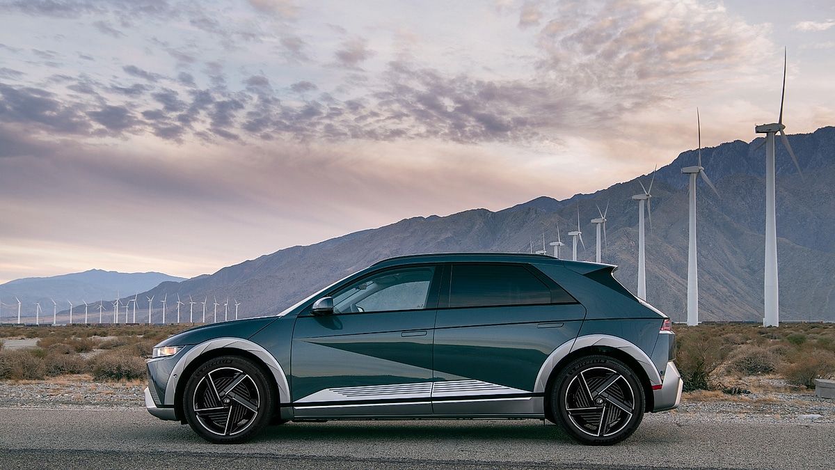 2024 Hyundai Ioniq 5 in deep teal with contrast silver trim shown in a side profile parked in a desert landscape surrounded by wind turbines and mountain ridges at sunset